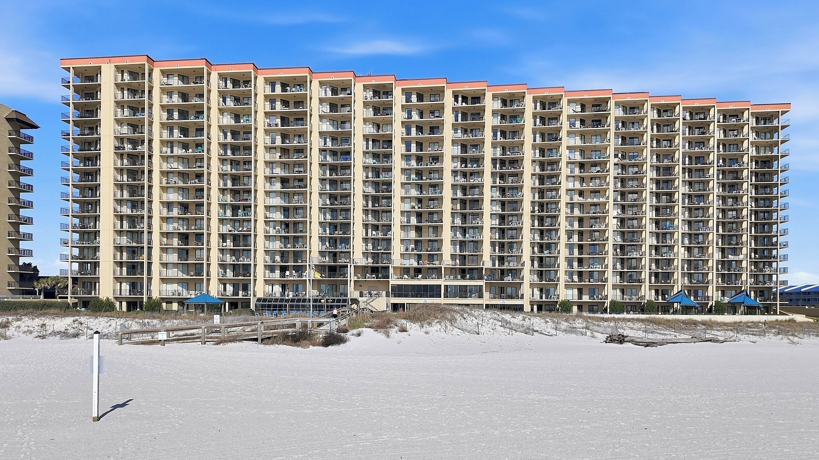 A high-rise hotel with multiple balconies, a flat roof, and a sandy beach in the foreground.