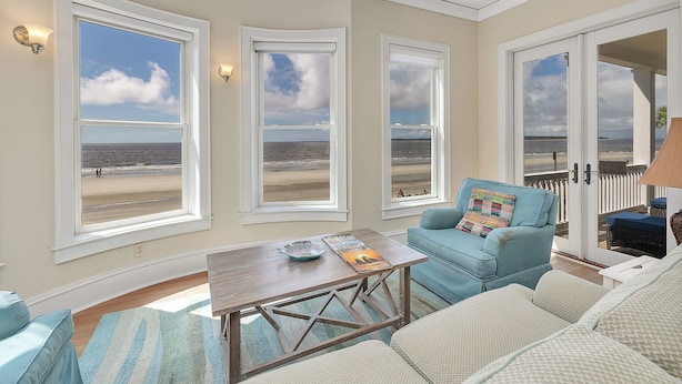 A spacious living room with large windows offering a view of the beach. There is a blue armchair and a wooden coffee table with magazines.