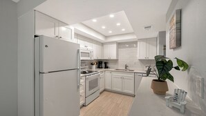 A modern kitchen with white cabinetry, a built-in oven, microwave, and dishwasher. There's a potted plant and a small table with chairs. - Siesta Key Beachfront Vacation Condo - great value and many amenities! (Sarasota)