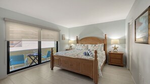 A bedroom with a wooden bed, two bedside tables with lamps, a large window with blinds, and a balcony with a table and chairs. - Siesta Key Beachfront Vacation Condo - great value and many amenities! (Sarasota)