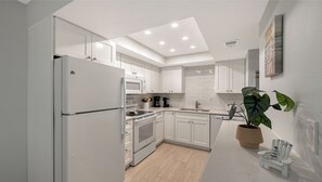 A modern kitchen with white cabinetry, a built-in oven, microwave, and dishwasher. There's a potted plant and a small table with chairs. - Siesta Key Beachfront Vacation Condo - great value and many amenities! (Sarasota)