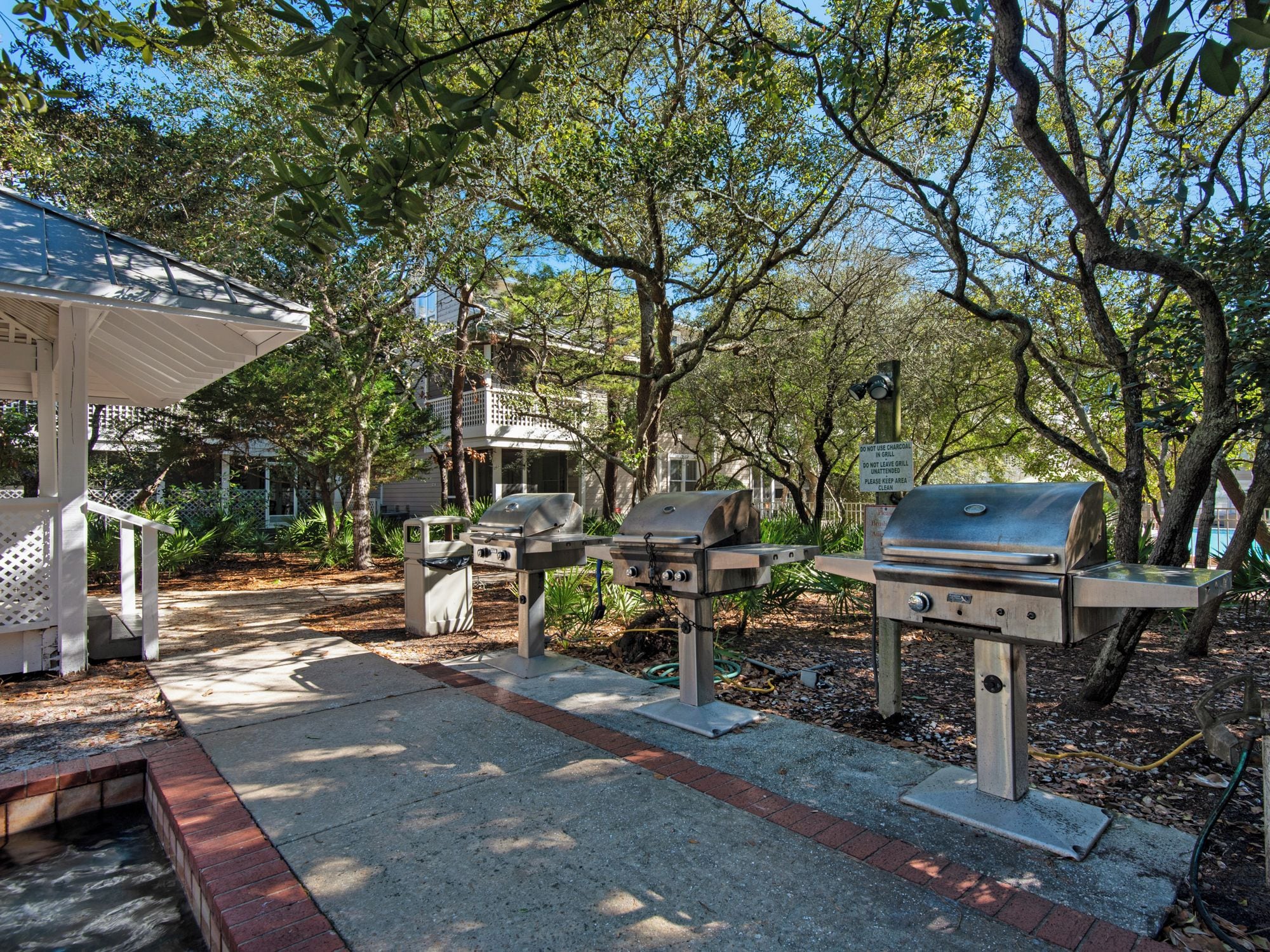A covered outdoor area with multiple barbecue grills, a paved walkway, and surrounding trees.
