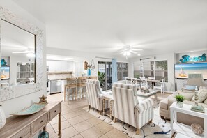 A spacious living area with a dining set, a kitchen in the background, and a large window with blinds.