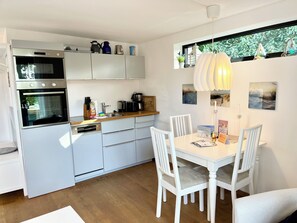 A modern kitchen with built-in appliances, a dining area with white chairs, and a view of greenery through a window. - WellenSpiel - unique atmosphere with rose garden (Wangerooge)