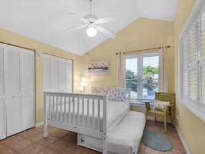 A bedroom with a white bed, a ceiling fan, a window with blinds, a chair, and a painting on the wall.