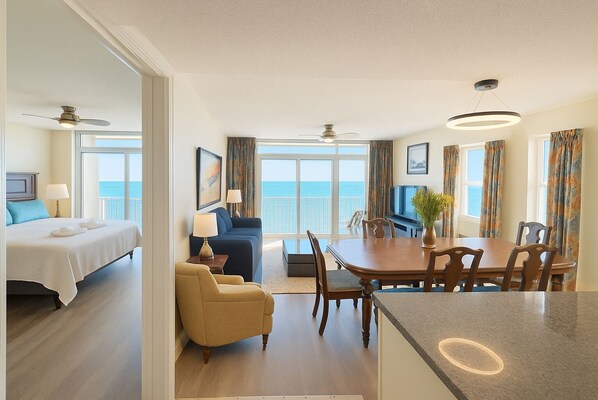 Ein Hotelzimmer mit Bett, einer Couch, einem Esstisch mit Stühlen, einem kleinen Sessel und einem Balkon mit Meerblick.