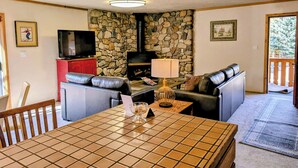 A living room with a stone wall, a fireplace, a television, a sofa, and a dining table. - Great 2 bed condo, Private deck & hot tub.  Best location in Ketchum! (Ketchum)
