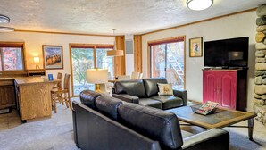 A cozy living area with a leather sofa, a wooden coffee table, and a TV on a cabinet. There is a kitchen area with a bar and a dining table. - Great 2 bed condo, Private deck & hot tub.  Best location in Ketchum! (Ketchum)