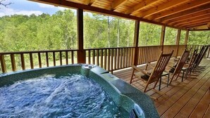 Un jacuzzi en una terraza de madera con sillas, rodeado por un barandal de madera, con vistas a un área boscosa.