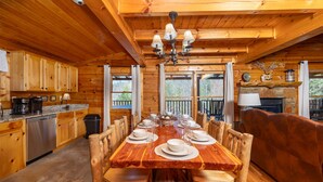 Interior de una cabaña de madera con una mesa de comedor preparada para seis personas, armarios de madera, una chimenea de piedra y una terraza de madera visible a través de las ventanas.