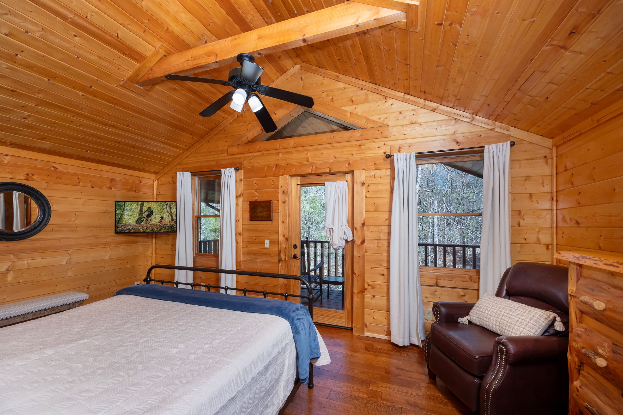 A wooden cabin room with a bed, a leather chair, a ceiling fan, and a balcony door.