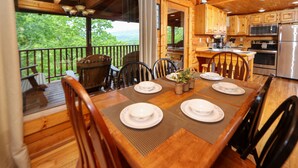A wooden dining table set with plates and chairs, a kitchen with wooden cabinets and appliances, and a view of greenery through a balcony railing. - Beautiful Mountain View Overlooking The Smokies! Hot Tub, Pool Table, Mini Golf (Sevierville)