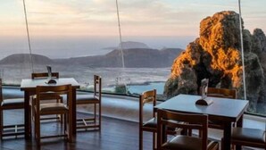 A dining area with tables and chairs, large windows with a view of a rocky coastline and distant mountains.