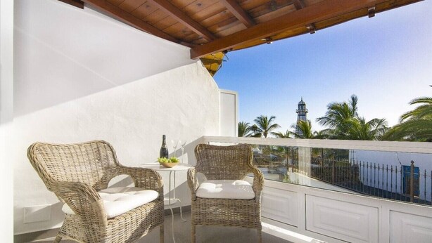 A balcony with wicker chairs and a small table, overlooking a body of water and palm trees.