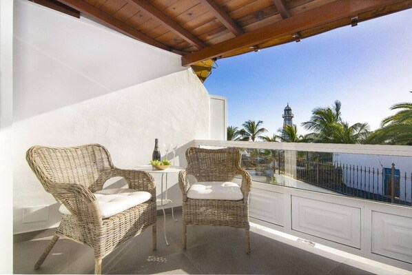 A balcony with wicker chairs and a small table, overlooking a body of water and palm trees.