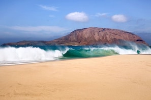 A beach with a large wave approaching the shore, a clear sky, and a mountain in the background.
