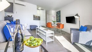 A modern living room with a sofa, a dining table with chairs, a flat-screen TV mounted on the wall, and an air conditioning unit.