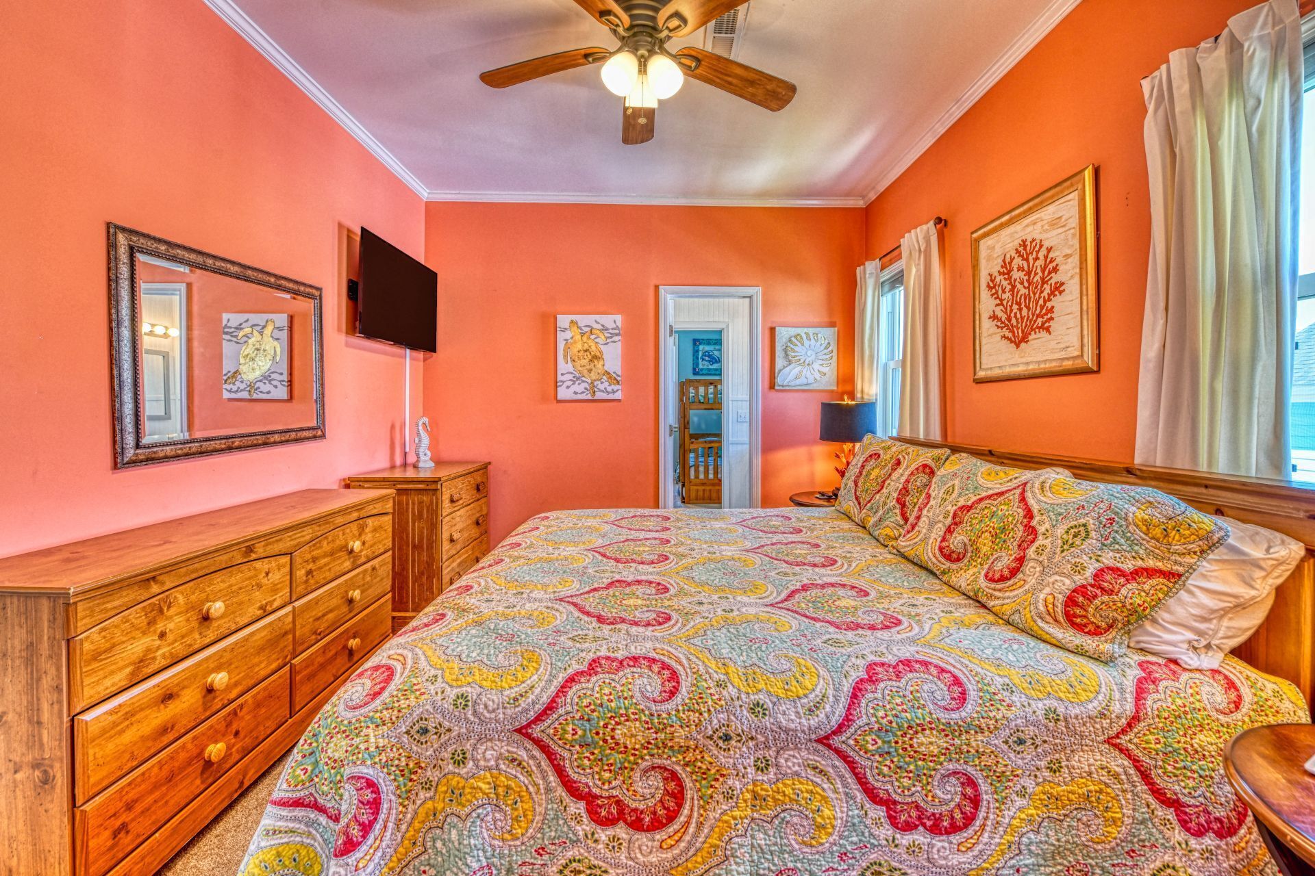 A bedroom with a patterned bedspread, wooden dresser, ceiling fan, and artwork on the walls.