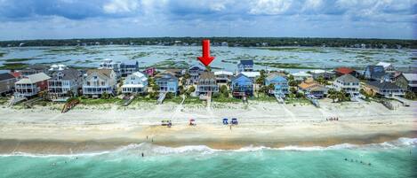 A coastal residential area with houses along the beach, a clear view of the ocean, and a red arrow pointing to a specific house.