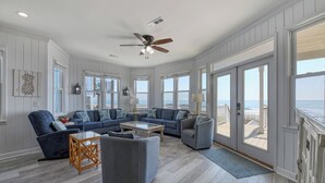 Een ruime woonkamer met een groot raam met uitzicht op het strand, een plafondventilator en een houten vloer.