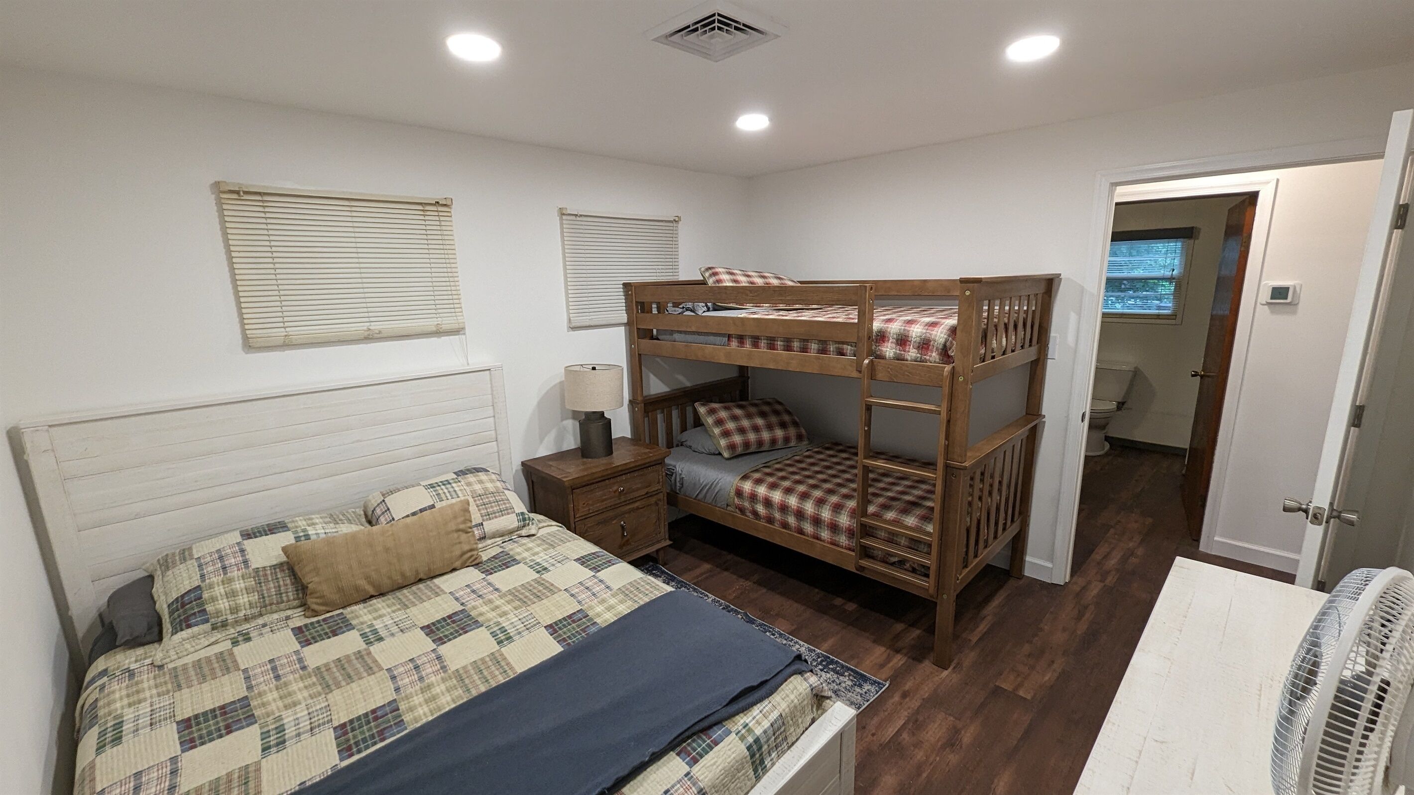 Une chambre avec un lit superposé, un lit simple, une table de chevet, une lampe et une fenêtre avec des stores.