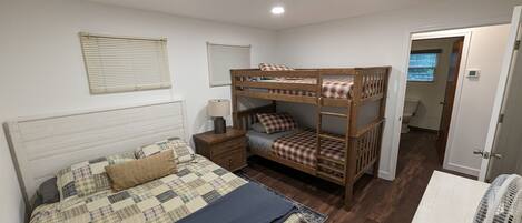 Une chambre avec un lit superposé, un lit simple, une table de chevet, une lampe et une fenêtre avec des stores.