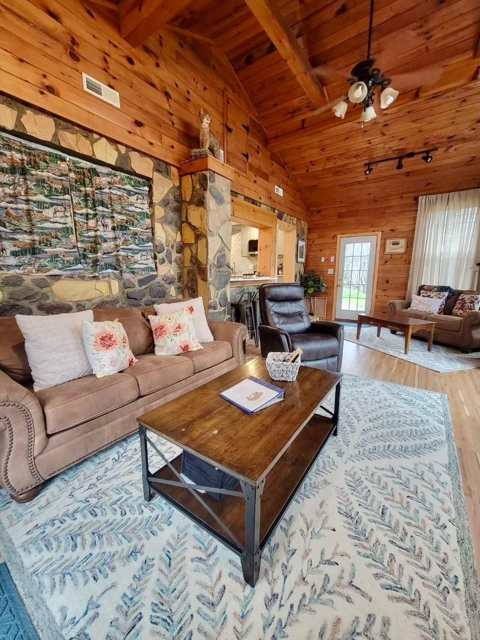A cozy cabin interior with a stone wall, wooden ceiling, a brown leather sofa, a coffee table, and a patterned rug.