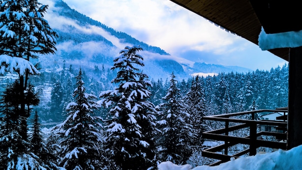 Paisaje nevado de montaña con una terraza de madera y vistas a árboles y montañas cubiertas de nieve.