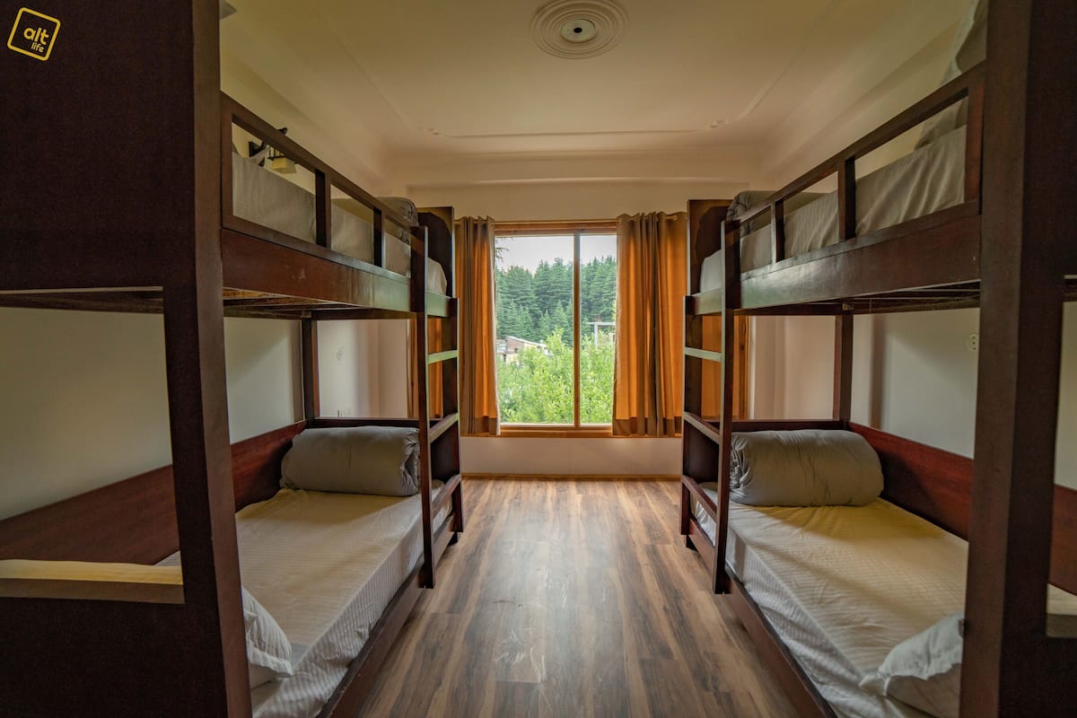 Quarto com beliches, janela com cortinas e vista para o verde lá fora.