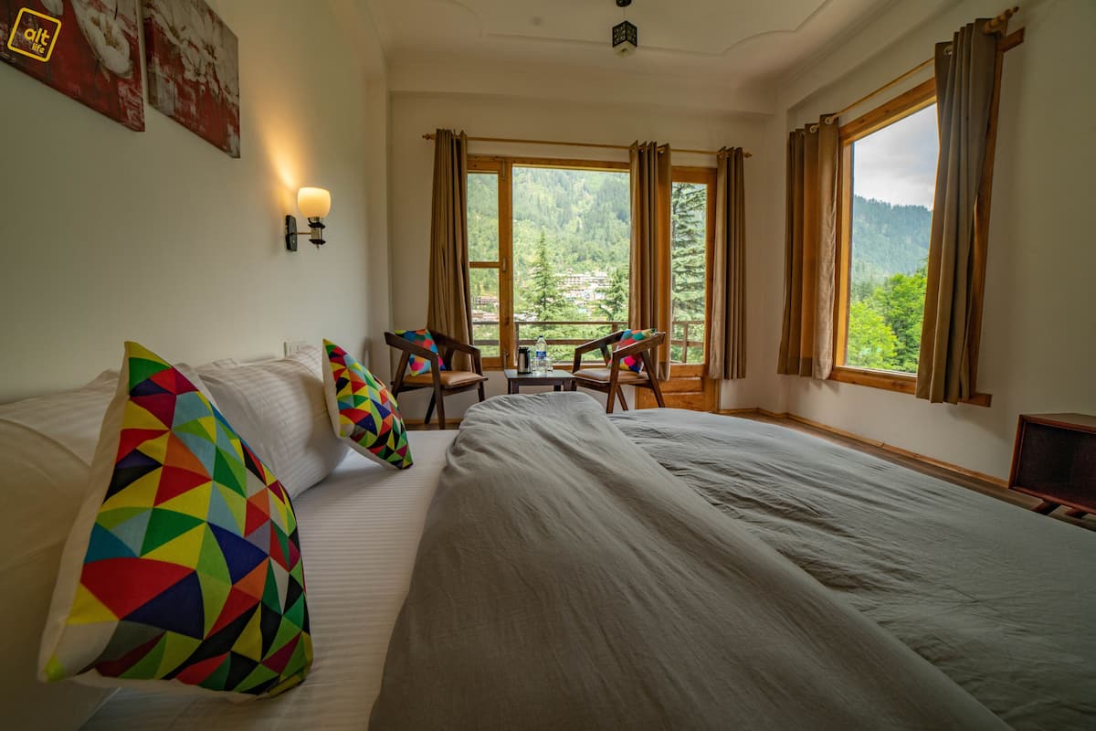 Quarto de hotel com uma cama grande, duas cadeiras, uma mesinha e uma janela com cortinas.