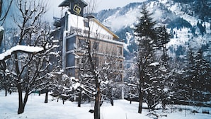 Un edificio cubierto de nieve con una torre destacada, rodeado de árboles cargados de nieve y montañas al fondo.
