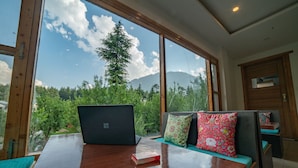 Una mesa de madera con una computadora portátil y un libro, una ventana con vista a árboles y montañas, y un banco con cojines.