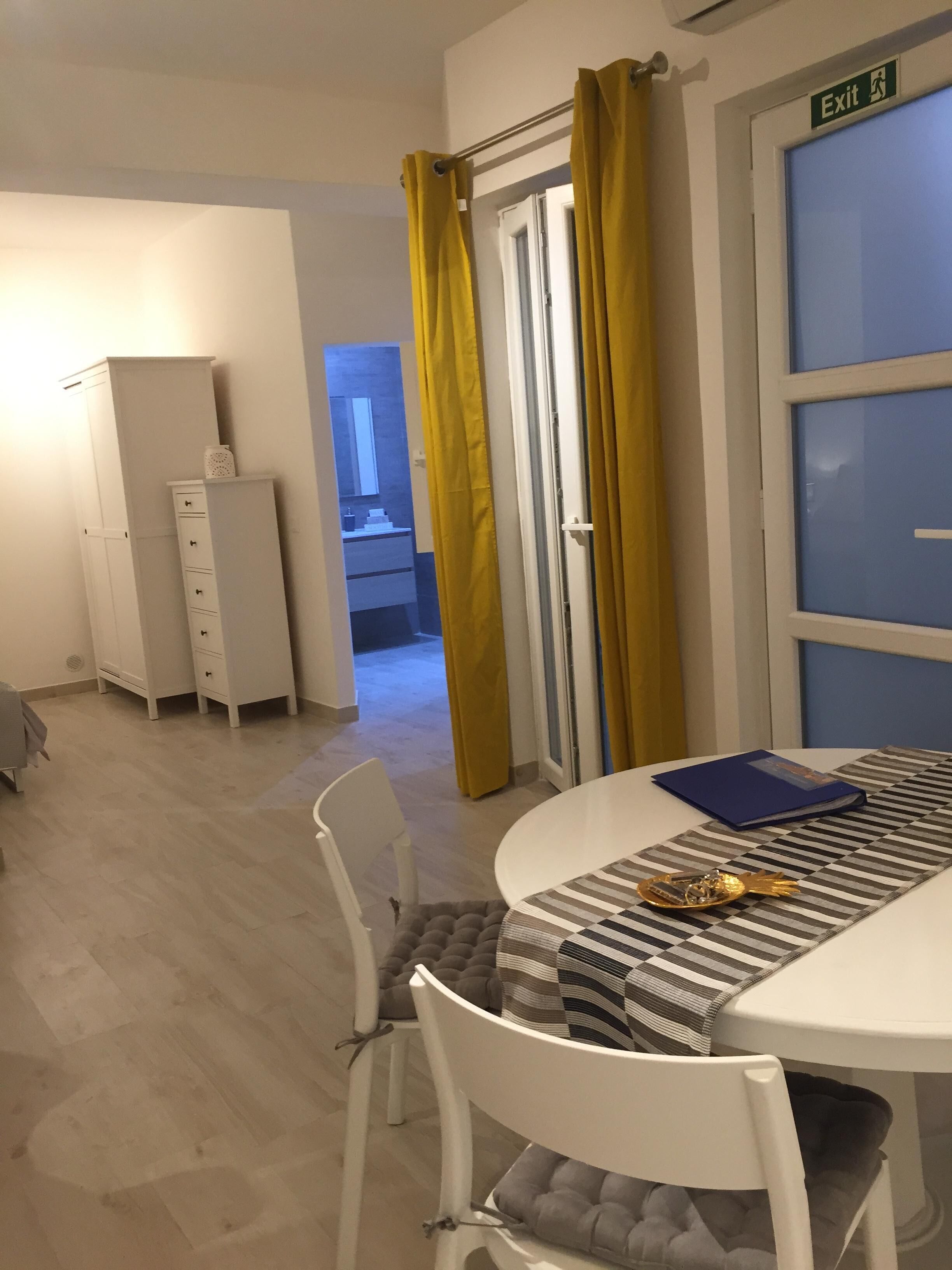 Una habitación con una mesa blanca, una carpeta azul y un adorno dorado. Hay una silla blanca con cojín gris, un armario blanco y una puerta con cortinas amarillas. Se observa un letrero de salida en la puerta.
