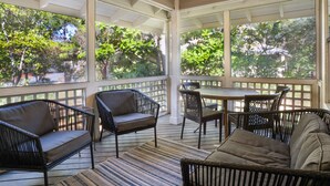 En overdækket terrasse med fletmøbler, et bord og store vinduer der giver udsigt til træer og en gade.