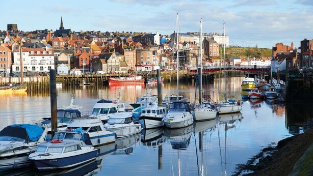 A harbor with boats docked, a waterfront promenade, and a town with historic buildings in the background.