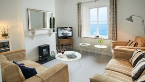 A cozy living room with a sofa set, a fireplace, a television, and a view of the sea through a window.