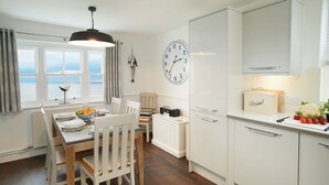 A modern kitchen with white cabinetry, a dining table with chairs, a clock on the wall, and a view of the sea through large windows.
