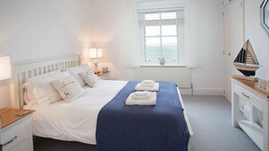 A neatly made bed with white linens and a blue accent, a bedside table with a lamp, a window with a view, and a decorative sailboat on a dresser.