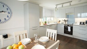 A modern kitchen with white cabinetry, a large wall clock, and a dining table set for four with a fruit bowl and glasses.