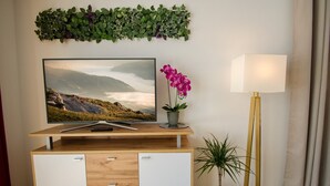 Ein modernes Wohnzimmer mit Fernseher, einem kleinen Schreibtisch, einer Topforchidee, einer hohen Stehlampe und einer grünen Wanddekoration mit Blättern.