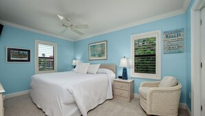 A bedroom with a bed, nightstands, a chair, a ceiling fan, and a beach-themed wall sign.