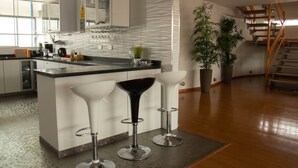 A modern kitchen with white cabinets, a marble countertop, and two bar stools. - APARTAMENTO PENTHOUSE  DE 350 M2, CON VISTA PANORAMICA DE LA CIUDAD. (Cusco)