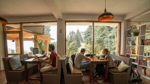 A modern living room with wooden flooring, built-in seating, large windows, and a bookshelf. - Alt Life - Dharamkot - Hostel (Dharamshala)