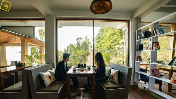 A cozy room with a large window, a wooden table, and a bookshelf. - Alt Life - Dharamkot - Hostel (Dharamshala)
