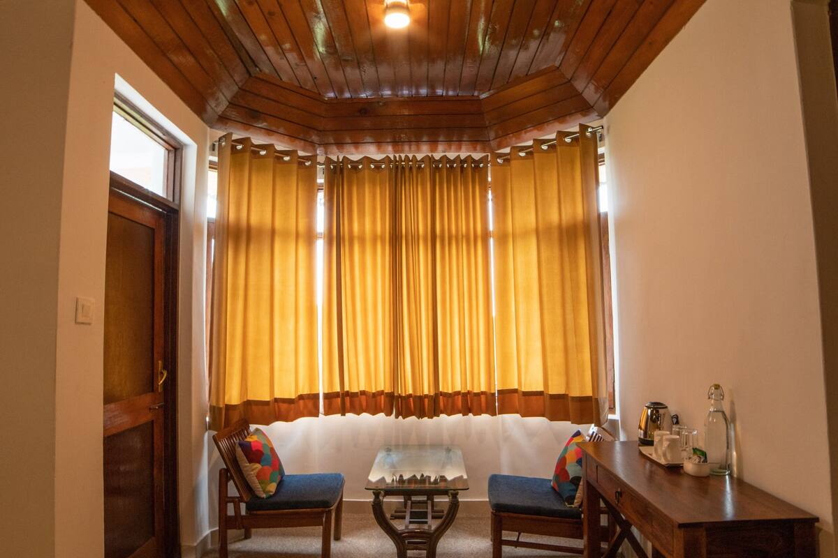 A room with a wooden ceiling, a window with yellow curtains, a sofa, two chairs, a small table, and a side table with a lamp.