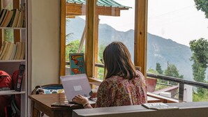 A person working at a wooden desk with a laptop, a bookshelf with books and a Superman poster, and a view of mountains through large windows. - Alt Life - Dharamkot - Hostel (Dharamshala)