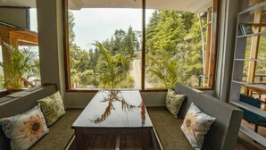 A room with a large window offering a view of a forested area and a dirt road. There is a wooden table with a vase of dried plants in the center. - Alt Life - Dharamkot - Hostel (Dharamshala)