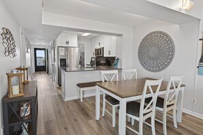 A modern kitchen with white cabinetry, a central island, and a dining area with a wooden table and white chairs. - CRC 1412 - Fourth Floor Condo with Beautiful Views (St Augustine)