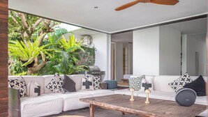 Un moderno salón con un sofá blanco, cojines estampados, una mesa de centro de madera y vistas a un follaje exuberante.