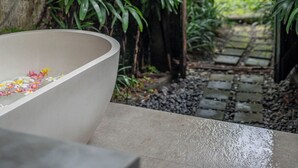 Una bañera de concreto con pétalos de flores, rodeada de vegetación exuberante y un sendero de piedra.